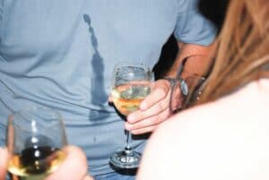 man holding glass of white wine with wine down his blue top